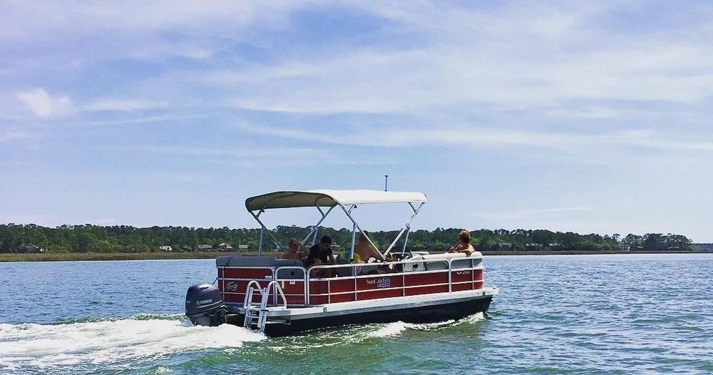 Red pontoon boat with canopy carrying people across a calm blue bay, leaving a gentle wake near a tree-lined shoreline under a sunny sky