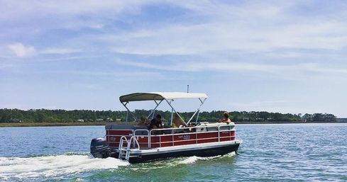 Red pontoon boat with canopy carrying people across a calm blue bay, leaving a gentle wake near a tree-lined shoreline under a sunny sky