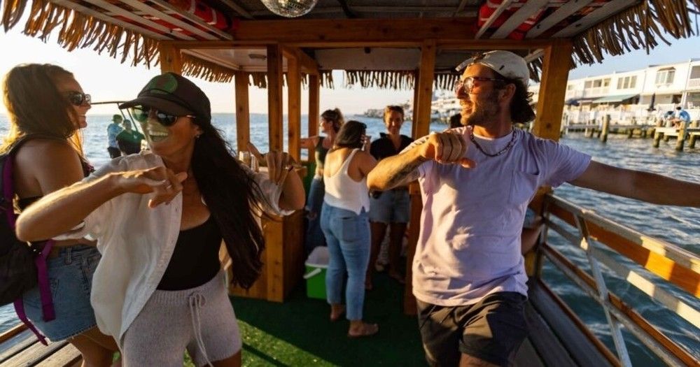 Group of friends dancing and laughing on a tiki-style party boat at sunset, cruising alongside a waterfront dock with piers and houses visible.