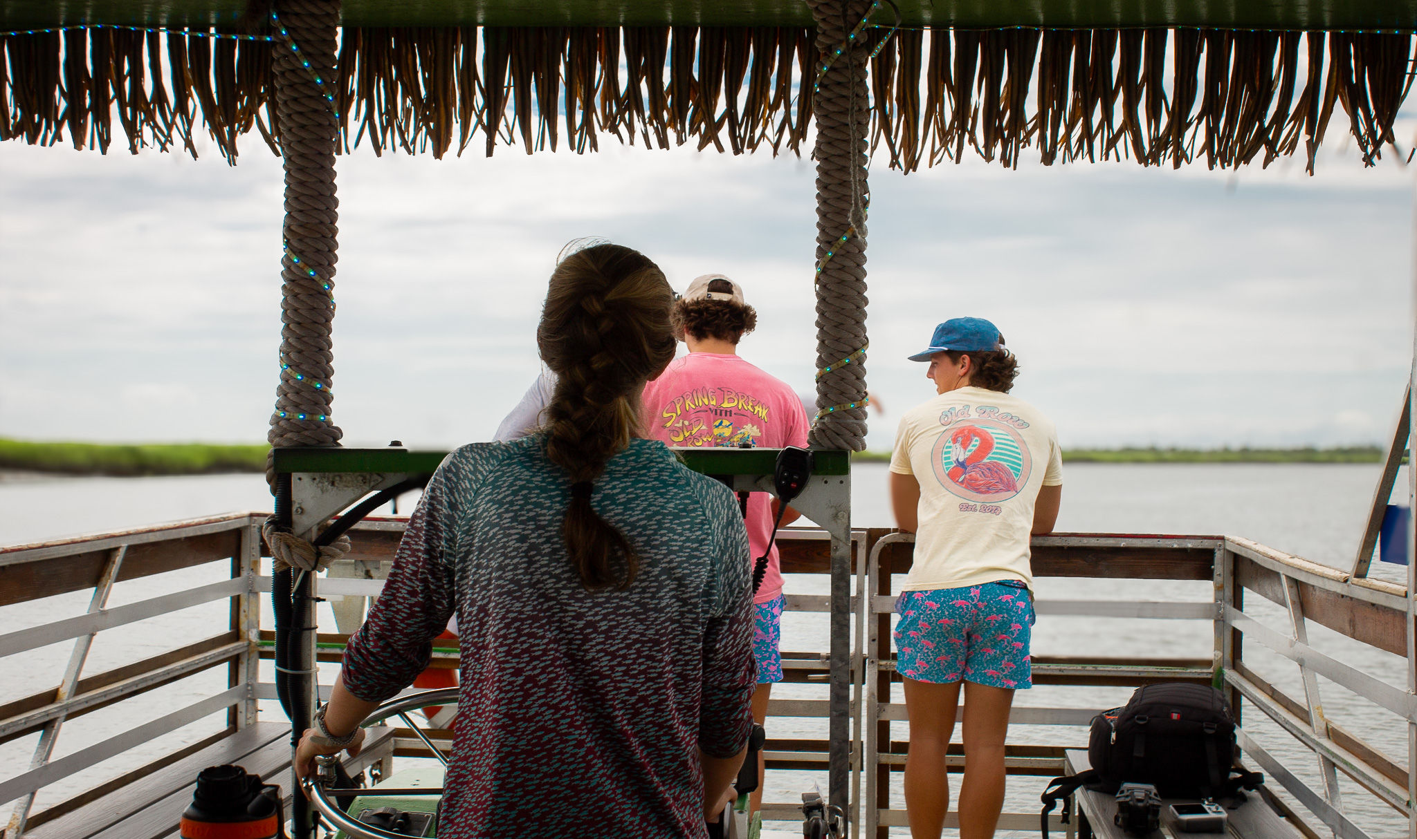 Three people on a small thatched-roof pontoon boat steering through coastal marsh waters under a cloudy sky