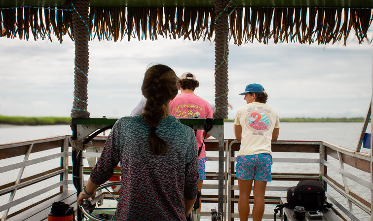 Three people on a small thatched-roof pontoon boat steering through coastal marsh waters under a cloudy sky