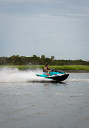 Person riding a turquoise jet ski across a calm coastal estuary, kicking up white spray past green marsh under a cloudy sky — fast water sports action.