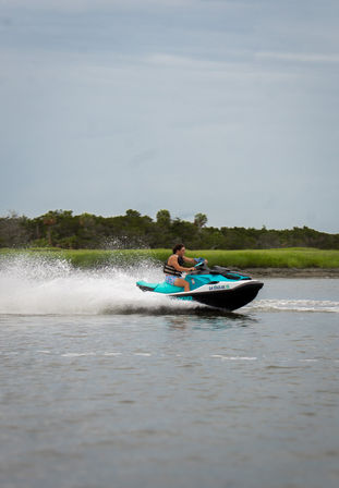 Person riding a turquoise jet ski across a calm coastal estuary, kicking up white spray past green marsh under a cloudy sky — fast water sports action.