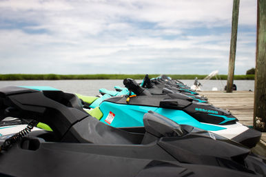Turquoise and black jet skis lined up on a wooden dock by a calm coastal marsh and bay under a partly cloudy sky
