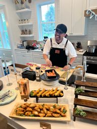 Happy chef in cap and apron preparing sushi at a bright white kitchen island, with trays of tempura shrimp and fried dumplings and a palm tree visible through the window.