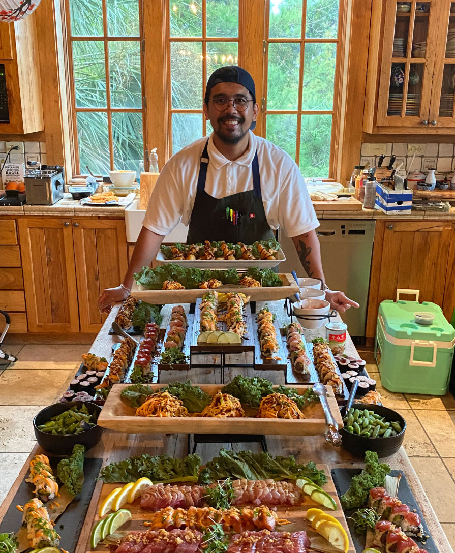 Smiling chef in a black apron and cap stands behind an abundant colorful sushi and sashimi spread on wooden boards in a sunlit rustic kitchen with large paned windows and greenery outside.