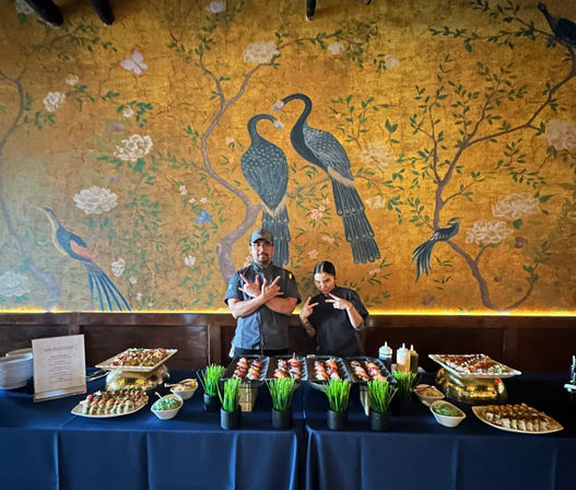 Two chefs playfully posing behind a sushi buffet with trays of nigiri, sauces and small grass planters, set against a large gold Asian-style mural of peacocks and flowering branches in a restaurant.