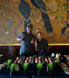 Two smiling sushi chefs pose behind trays of nigiri sushi on a blue-clothed table, with decorative gold bird mural on the wall — cozy sushi bar scene.