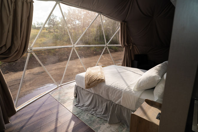 Sunlit glamping geodesic dome interior with a cozy white bed and folded blanket, panoramic triangular windows overlooking a dirt road and grassy countryside.