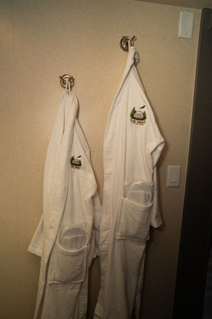Two white plush bathrobes with embroidered nature logo and packaged slippers tucked in pockets, hanging on chrome hooks in a hotel bathroom with beige walls