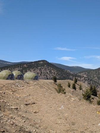 Three pale-green geodesic domes dot a rocky mountain ridge with scattered pine trees, a dry dirt slope and layered forested hills beneath a clear blue sky