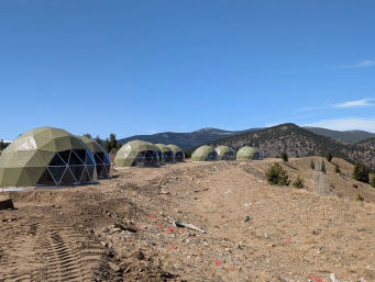 Row of olive-green geodesic glamping domes on concrete pads along a cleared mountain ridge with tire tracks, scattered rocks, pine trees and forested peaks under a clear blue sky.