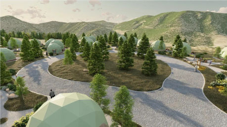 Aerial view of green geodesic glamping domes nestled among pine trees with winding gravel paths in a sunlit mountain valley.