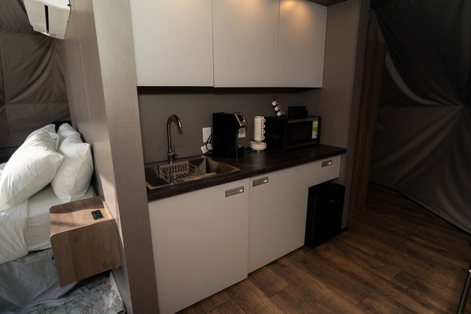 Compact modern kitchenette in a cozy studio suite with a stainless sink and faucet, single-serve coffee maker and mug carousel, microwave, mini-fridge, overhead cabinets, and an adjacent bed with white pillows on wood-look flooring.