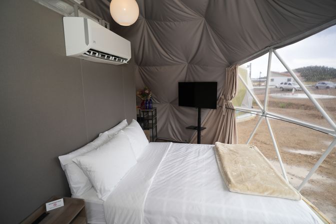 Cozy modern glamping geodesic dome interior with white-made bed, wall air conditioner, flat-screen TV and panoramic dome window overlooking a campsite.
