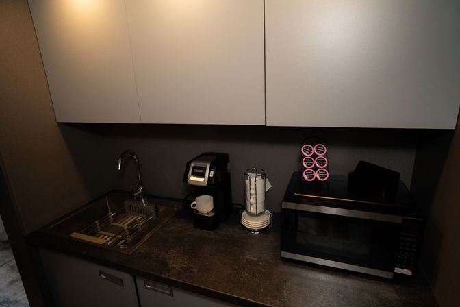 Compact modern kitchenette with stainless sink and faucet, single-serve coffee maker with white mug, glass jar of white pods, a rack of coffee pods, and black microwave beneath gray cabinets.