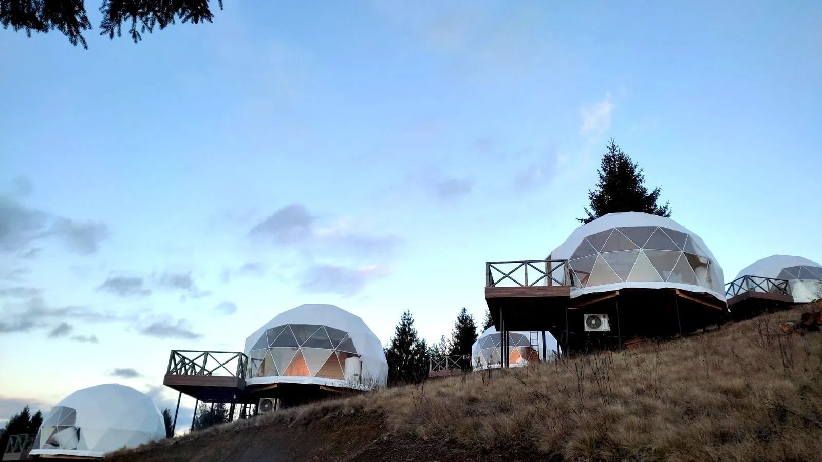 Cozy white geodesic glamping domes with wooden decks on a grassy mountain hillside at dusk, pine trees and pastel sky in the background