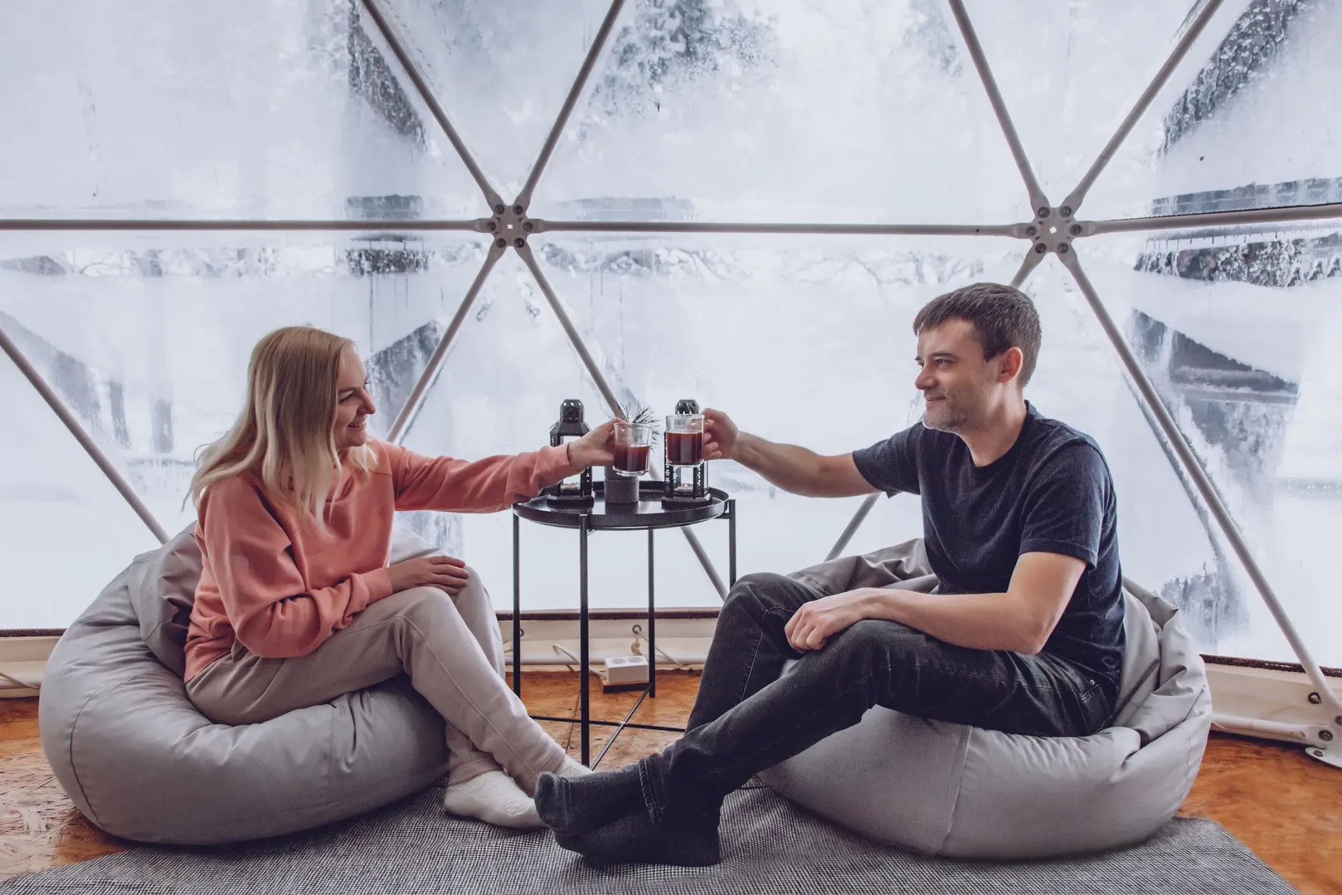 Couple clinking steaming mugs inside a frosted geodesic dome, seated on gray beanbag chairs around a small table — cozy winter glamping retreat with snowy windows.