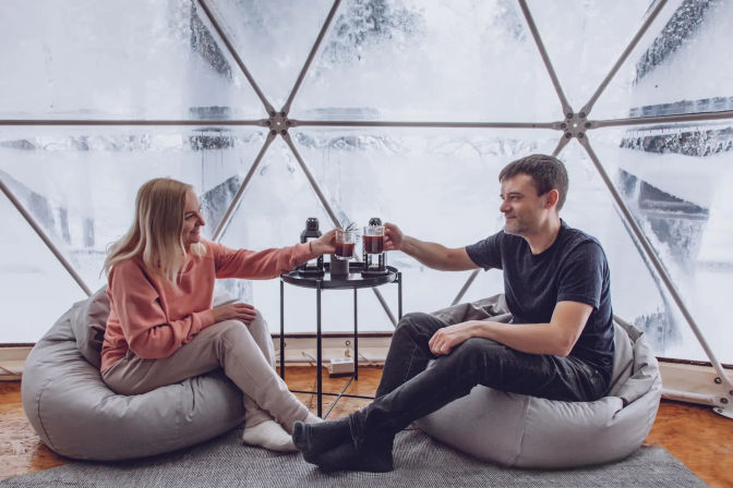 Couple clinking steaming mugs inside a frosted geodesic dome, seated on gray beanbag chairs around a small table — cozy winter glamping retreat with snowy windows.