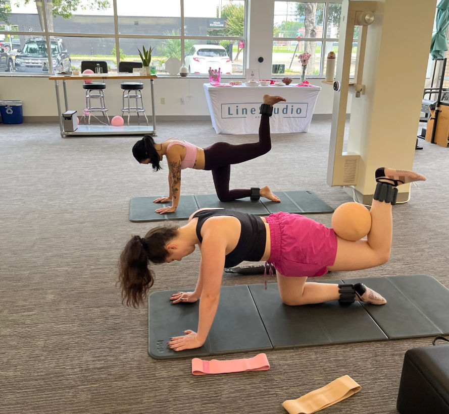 Two women in sporty gear doing pilates-style glute kickbacks on mats in a bright indoor fitness studio, using ankle weights and a small exercise ball with resistance bands nearby.