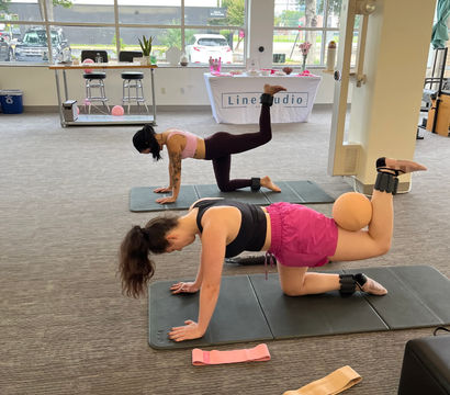 Two women in sporty gear doing pilates-style glute kickbacks on mats in a bright indoor fitness studio, using ankle weights and a small exercise ball with resistance bands nearby.