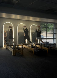 Three people doing Pilates reformer exercises holding medicine balls overhead in a dim fitness studio with backlit arched mirrors and a large window