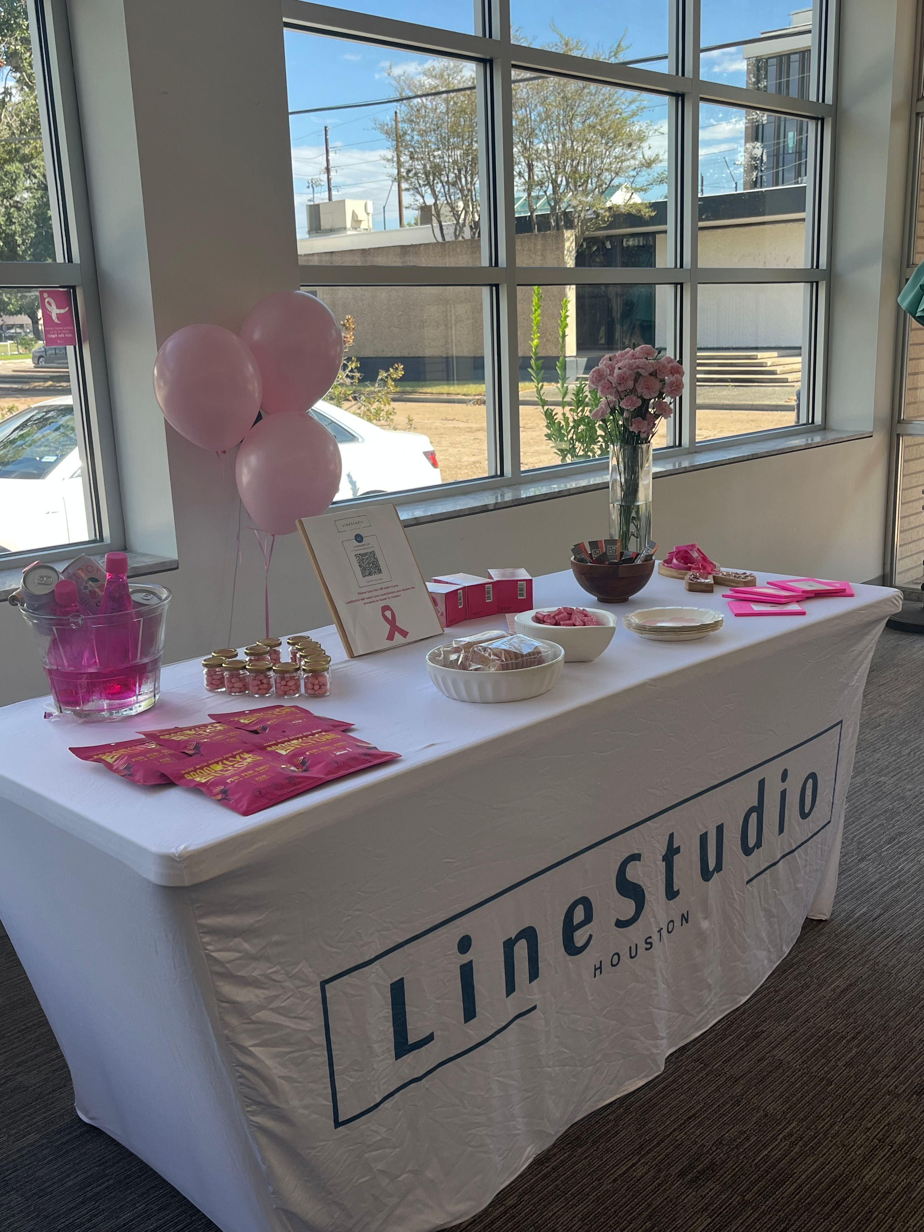 Houston office event table with pink balloons, a pink-ribbon sign, bouquet of pink roses and pink-themed snacks and giveaways on a white-covered table by large windows