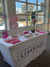 Houston office event table with pink balloons, a pink-ribbon sign, bouquet of pink roses and pink-themed snacks and giveaways on a white-covered table by large windows