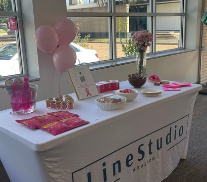 Houston office event table with pink balloons, a pink-ribbon sign, bouquet of pink roses and pink-themed snacks and giveaways on a white-covered table by large windows