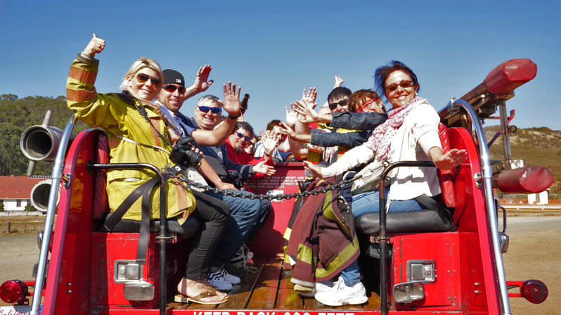 Cheerful group of adults and children waving from the open rear seats of a red fire engine on a sunny outdoor sightseeing ride, with sunglasses, a firefighter jacket and cameras visible.