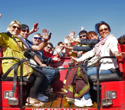 Cheerful group of adults and children waving from the open rear seats of a red fire engine on a sunny outdoor sightseeing ride, with sunglasses, a firefighter jacket and cameras visible.