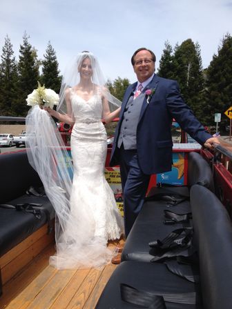 Smiling bride in a lace gown and long veil holding white bouquet stands beside a man in a navy suit with a pink tie on an open-top red vehicle with wooden floor during an outdoor wedding with tall evergreen trees behind them