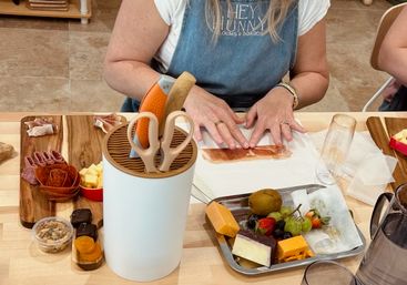 Busy hands assembling prosciutto at a kitchen table for a charcuterie board, surrounded by cheeses, grapes and strawberries on wooden boards, a metal serving tray, and a utensil caddy with scissors and knives.