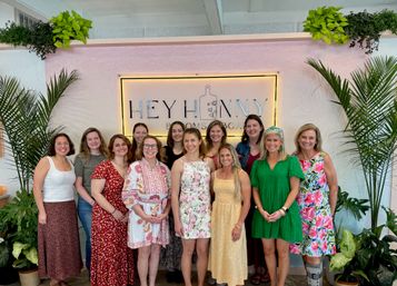 Group of smiling women posing indoors in front of a pink decorative sign framed by lush green plants, wearing colorful floral and summer dresses.