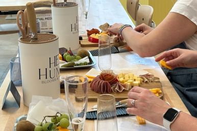 Hands-on charcuterie workshop: people assembling wooden cheese boards with cured meats, cubed cheeses, grapes, kiwi and champagne flutes on a bright indoor table.