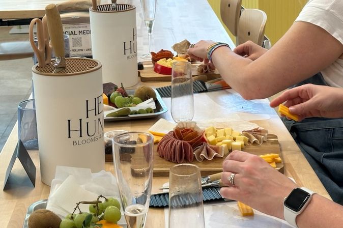 Hands-on charcuterie workshop: people assembling wooden cheese boards with cured meats, cubed cheeses, grapes, kiwi and champagne flutes on a bright indoor table.