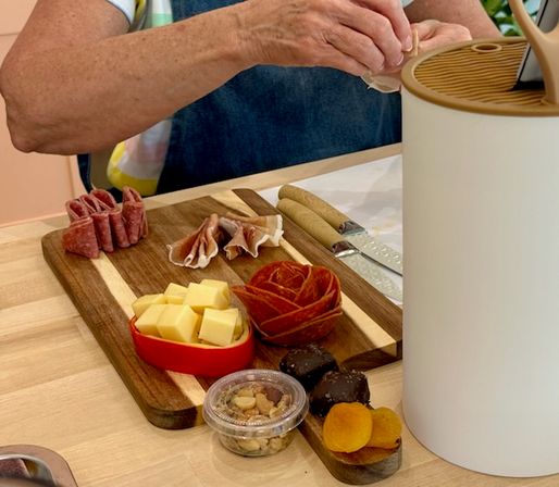 Hands arranging a wooden charcuterie board on a kitchen countertop with cubed cheese, rolled salami, folded prosciutto, a pepperoni rose, dried apricots, chocolate truffles and a small cup of mixed nuts.