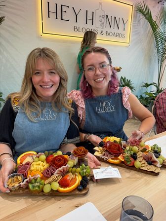 Two smiling women in aprons holding decorative charcuterie grazing boards with fresh fruit, cheeses, cured-meat roses, crackers and nuts at an indoor hands-on charcuterie workshop with plants and a lit sign in the background.
