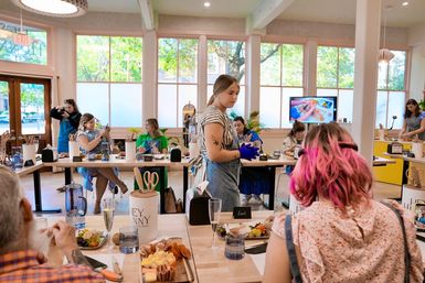 Vibrant daytime charcuterie workshop in a bright studio with large windows — instructor in denim apron and purple gloves demonstrates while participants at communal tables assemble cheese and snack boards.