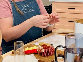 Hands crafting a colorful charcuterie board on a wooden board with folded salami, cubed cheeses and crackers on a kitchen countertop, person wearing a denim apron arranging the meats.