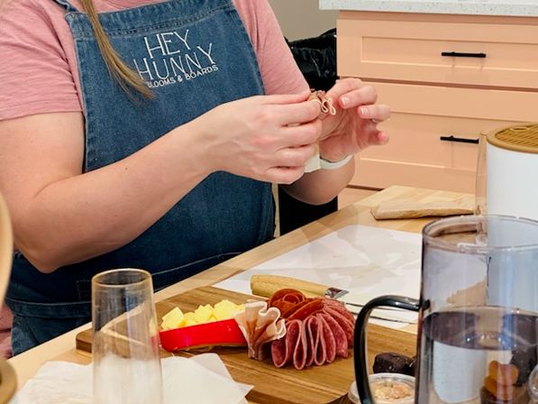 Hands crafting a colorful charcuterie board on a wooden board with folded salami, cubed cheeses and crackers on a kitchen countertop, person wearing a denim apron arranging the meats.