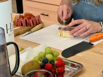 Hands slicing a block of cheese on parchment at a home kitchen counter, with a wooden charcuterie board of folded salami and a stainless tray of grapes, strawberries, blueberries and kiwi in the foreground.