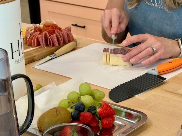 Hands slicing a block of cheese on parchment at a home kitchen counter, with a wooden charcuterie board of folded salami and a stainless tray of grapes, strawberries, blueberries and kiwi in the foreground.