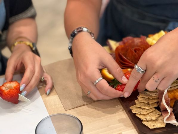 Close-up of two hands arranging strawberries on a charcuterie board with crackers, prosciutto, orange slices and a cured-meat rose on a light wooden table.