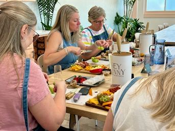 Four participants at a sunlit indoor workshop assembling colorful charcuterie boards with cheeses, fruit, nuts and crackers on a wooden table.