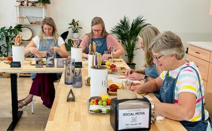 Four women in aprons making DIY charcuterie boards at long wooden tables in a bright, plant-filled craft studio with fruit, cheese, knives and communal supplies.