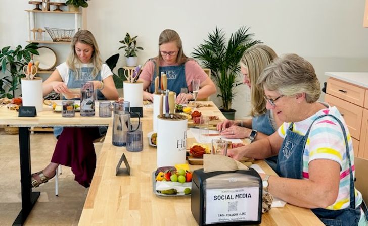 Four women in aprons making DIY charcuterie boards at long wooden tables in a bright, plant-filled craft studio with fruit, cheese, knives and communal supplies.