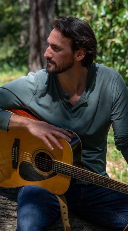 Relaxed man sitting on a log in a sunlit woodland, holding an acoustic guitar across his lap — outdoor nature portrait of a musician in a forest clearing.