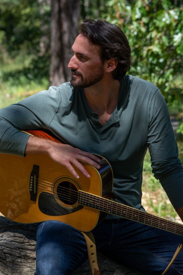 Relaxed man sitting on a log in a sunlit woodland, holding an acoustic guitar across his lap — outdoor nature portrait of a musician in a forest clearing.