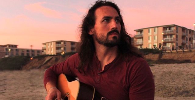 Long-haired bearded man in a burgundy shirt playing an acoustic guitar on a sandy beach at sunset with seaside apartment buildings in the background